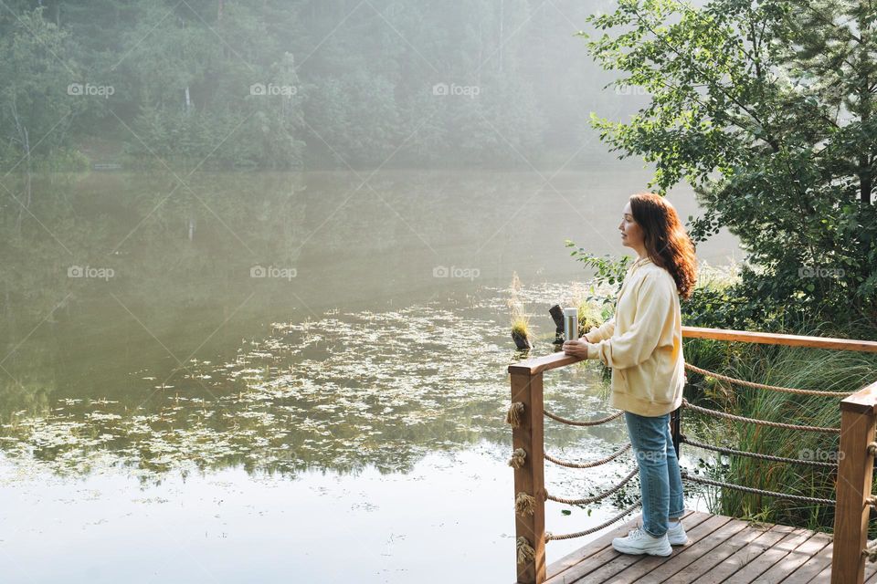 Brunette woman in yellow hoodie with thermo mug relaxing near lake in early morning