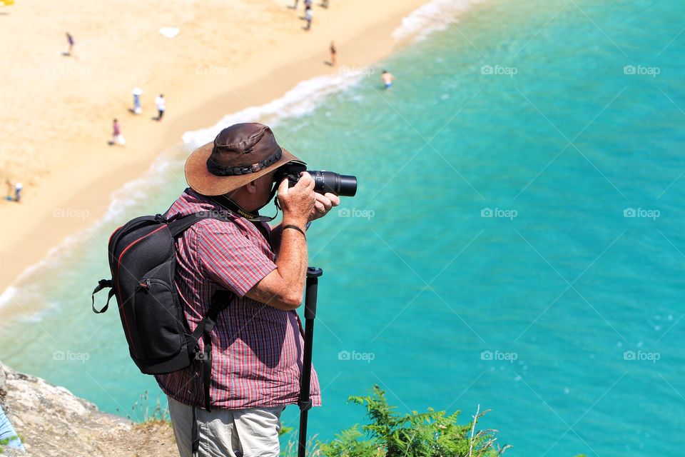 An elderly man taking photographs whilst overlooking a sandy beach and green ocean in Cornwall, UK.
