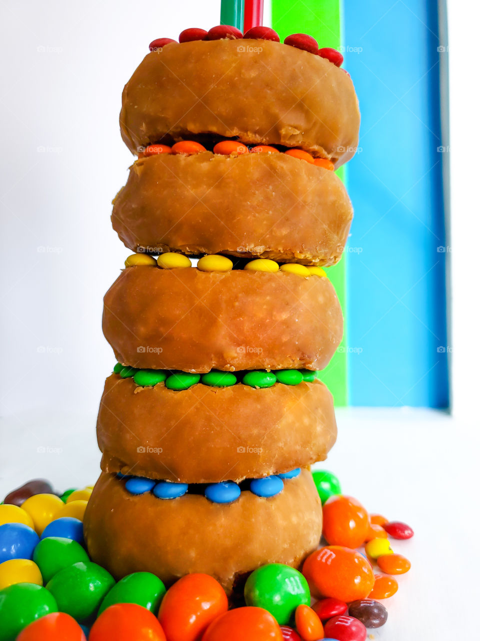 Five stacked donuts decorated with M&M minis and surrounded by peanut M&Ms on a white surface with a blue, green and white solid background. Reminiscent of the classic Fisher Price rock-a-stack infant toy.