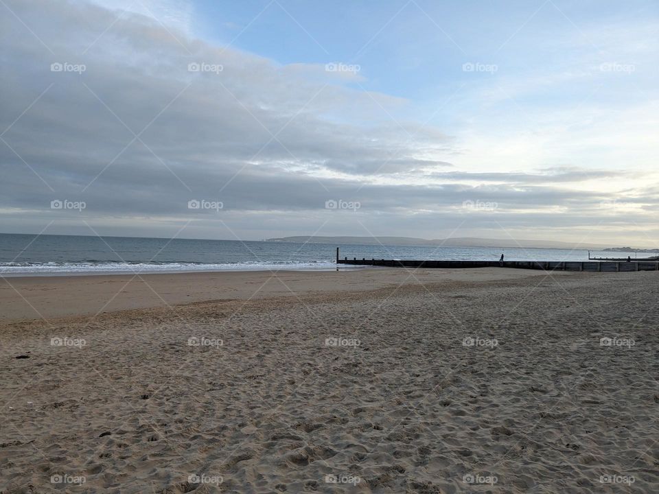 An empty stretch of sandy beach in the early evening. Prints from earlier activity visible in the sand. Sun mostly hidden behind clouds