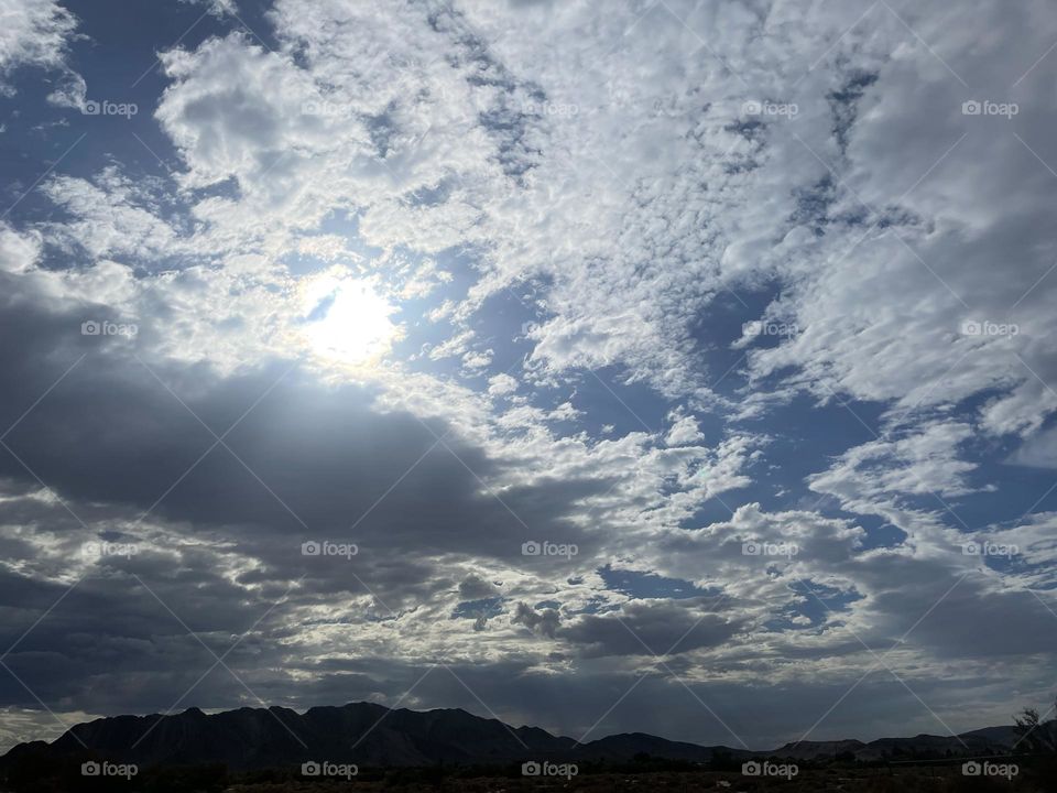A cloudy sky with the sun peeking through and mountains below the clouds.