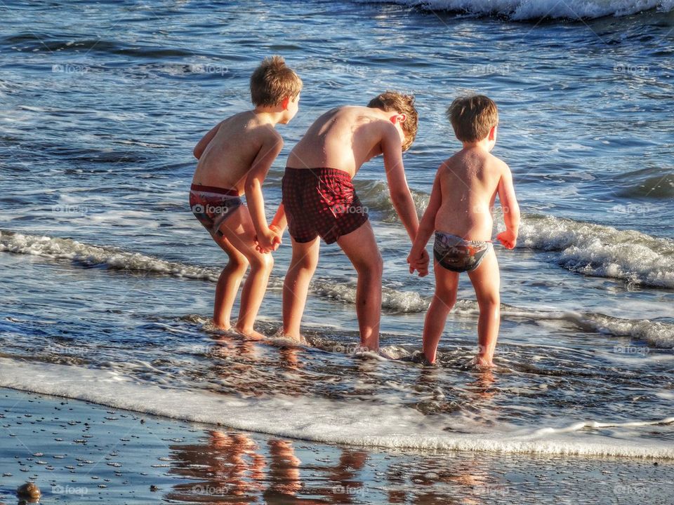 Three Young Brothers Playing In Ocean Waves. Brothers Holding Hands On The Beach
