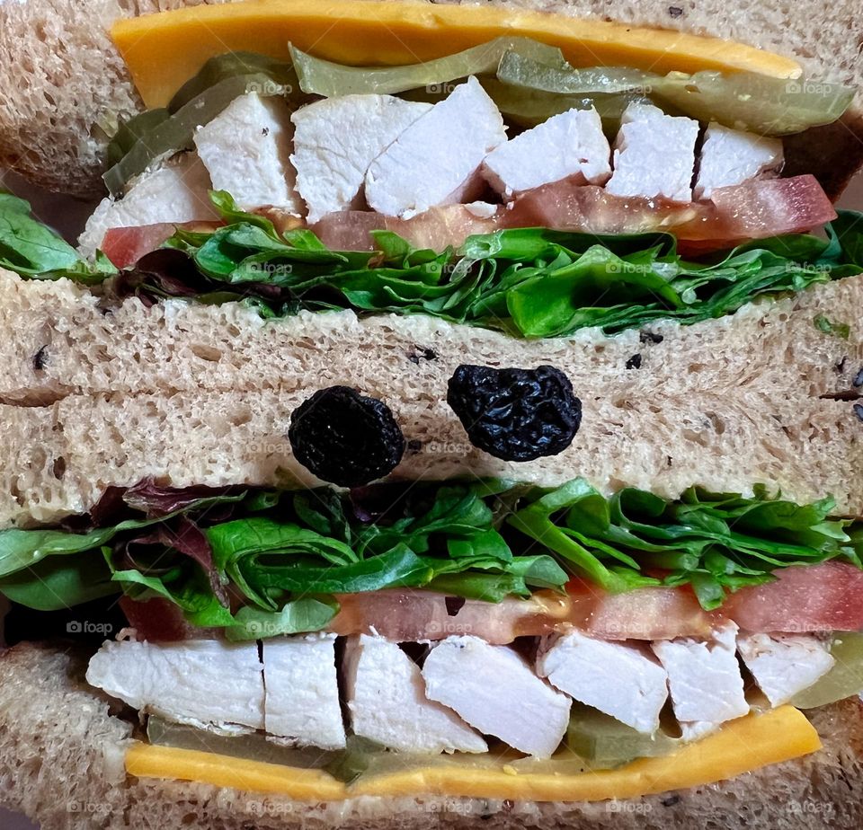 Chicken sandwich for lunch with tomatoes, lettuce and cheese. With two raisins as decorations. And now it look like a doubled teeth upper and lower.