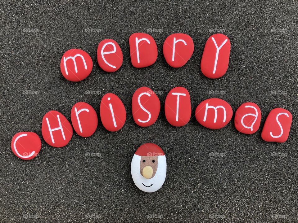 Merry Christmas text with red colored stones, Santa Claus face on a stone over black volcanic sand