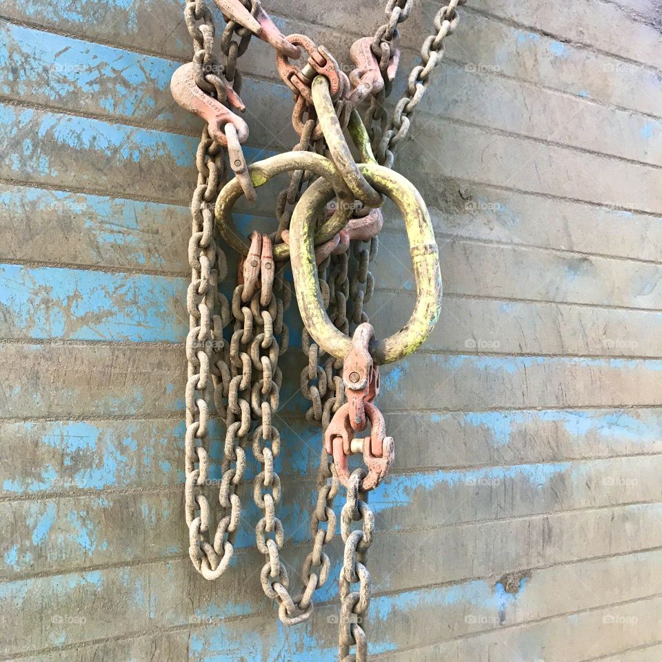Strong used chains with hooks and connections hanging from metal surface/wall with remnants of blue paint.