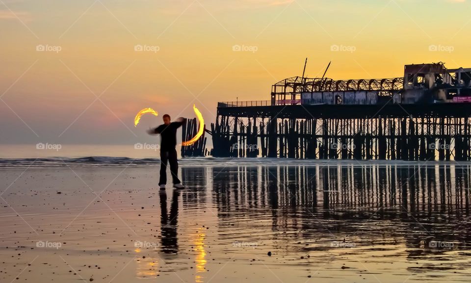 A man spins fire poi on the beach in front of the burnt out remains of the old Hastings pier