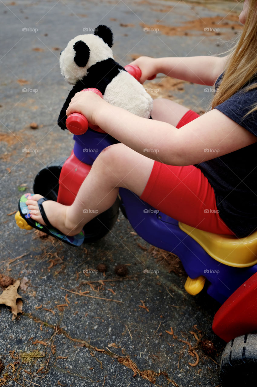A young girl with long blonde hair in shorts and sandals zooming along on her low-riding tricycle with her stuffed panda bear riding on the handle bars. 
