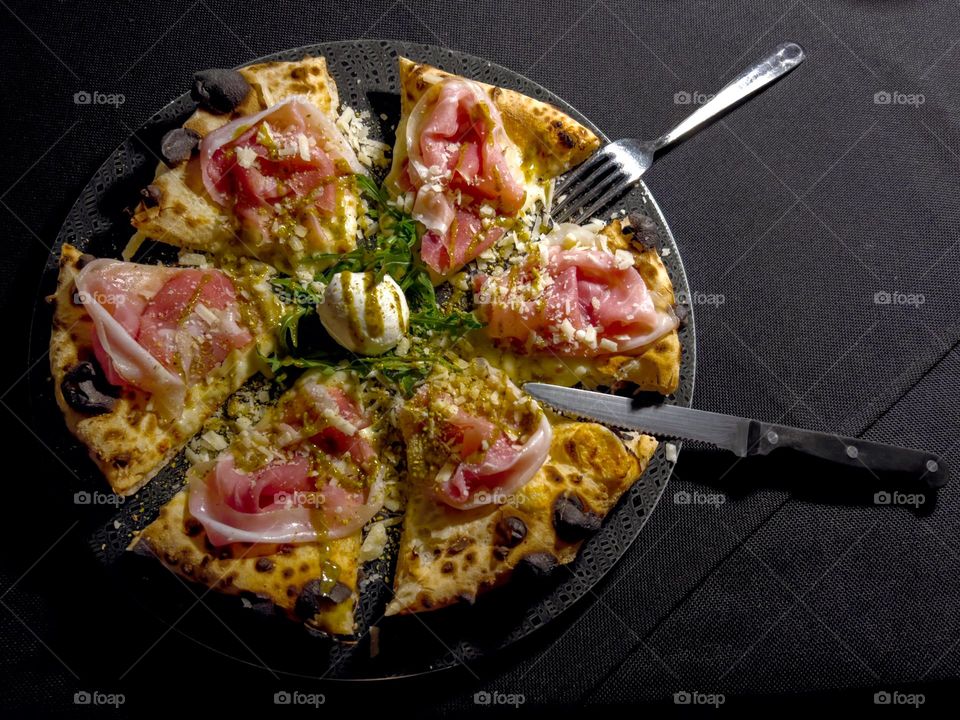 Italian pizza with ham, mozzarella, ruccola as well as knife and fork pointing to the mozzarella in the middle.