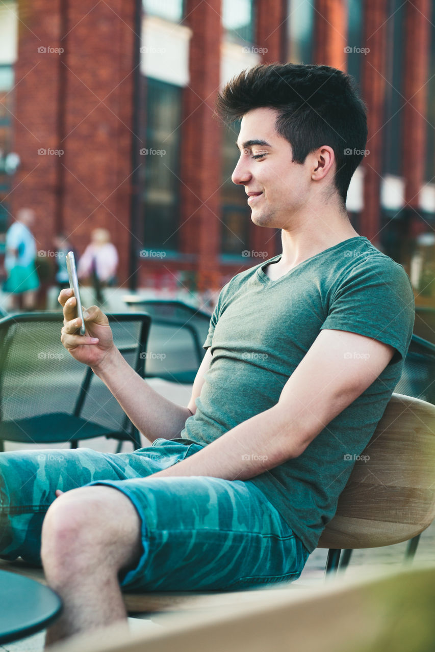 Young man having fun with smartphone, reading funny texts, sitting in center of town