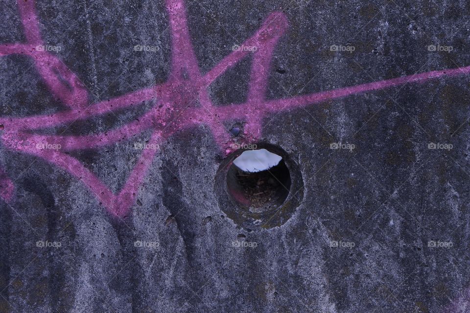 A closeup of a hole in a grey concrete jersey barrier with a pink painted tag symbol. 