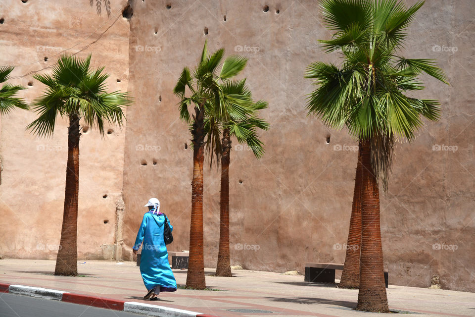 Moslim woman is walking before a city wall with palm trees in Marocco