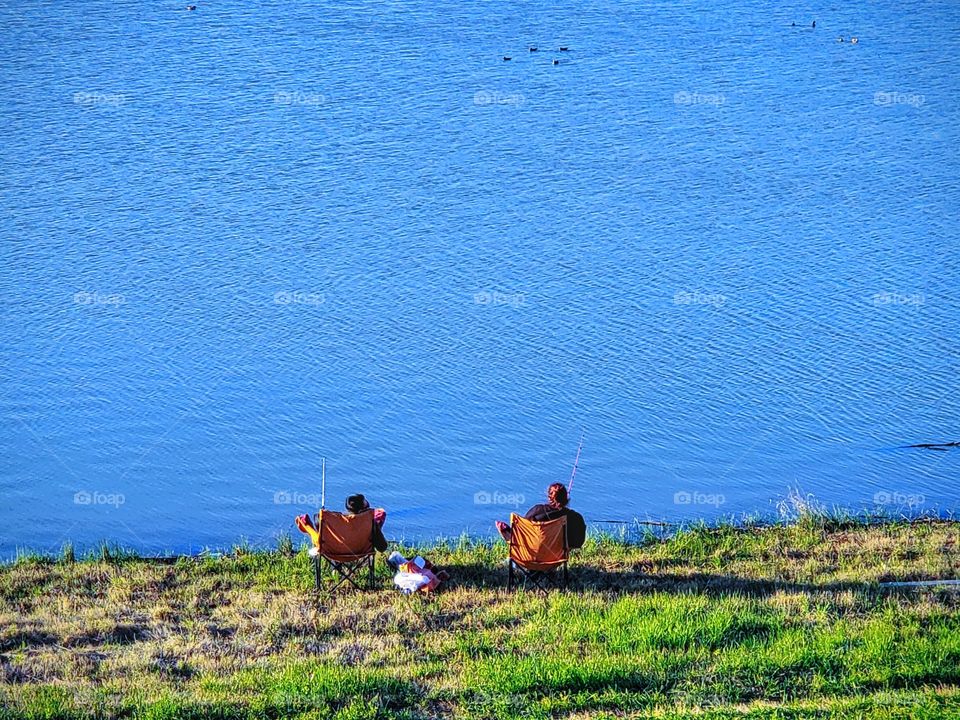 Landscape view of two adults relaxing sitting on an orange chairs, on green grass,  fishing while facing the lake.