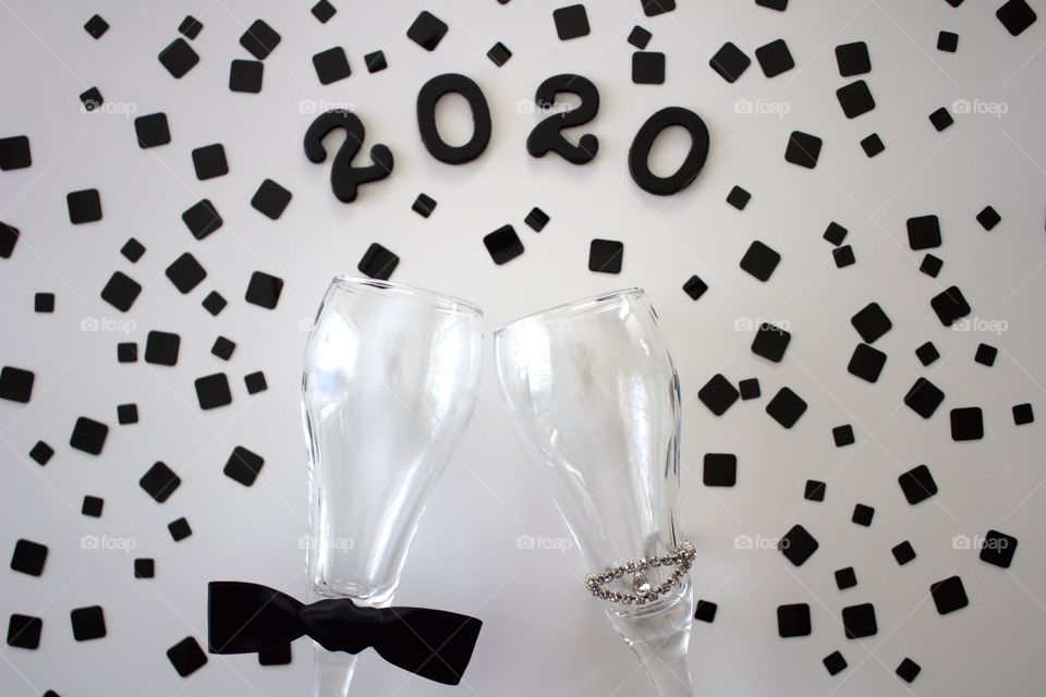 Conceptual overhead view of champagne glasses representing a man and a woman with the year ‘2020’ in black with black confetti on a white background - landscape format