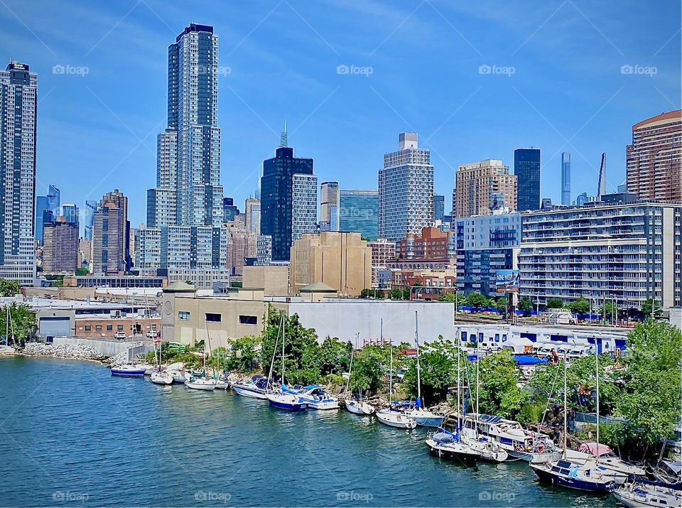 A view of the “E River” at “Newtown Creek” on the same glorious summer day. Not a cloud in the sky. The trees on the shore lush & green, the boats bright & white and the NYC skyline looks like elements of a cubist painting. 2022. Hypnotic Productions