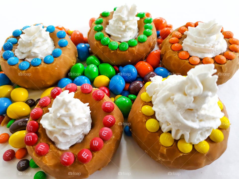 Donuts decorated with M&M minis, topped off with whip cream and surrounded by peanut M&Ms on a white surface.