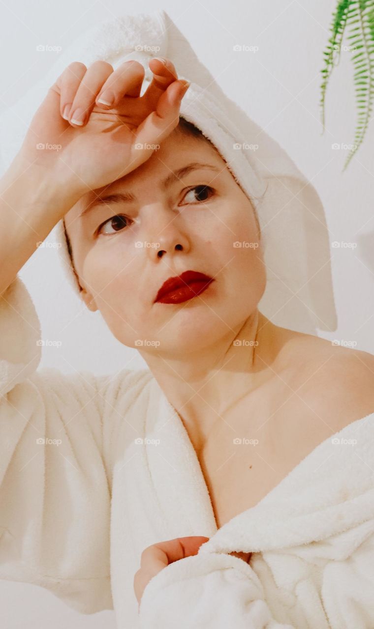 Portrait of one young beautiful Caucasian brunette girl with red lips in a white bathrobe with a towel on her head, looking to the side, opening one shoulder with her hand, close-up side view.