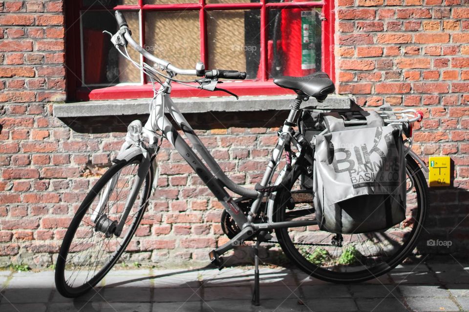 A gray two-wheeled bicycle with a dark gray bag on the rear of the trunk stands parked near a house with a red brick wall and a window with iron bars on a street in the city of Bruges on a clear sunny day, close-up side view.