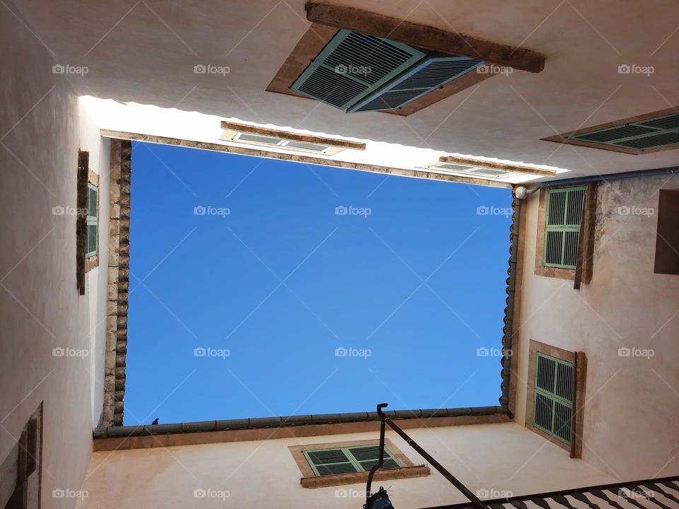 View from the ground up of a courtyard at a Mediterranean traditional ancient Manor. Blue sky can be seen above (at the center of the image). White walls and green Majorcan blinds  Calvia, Majorca, Spain.