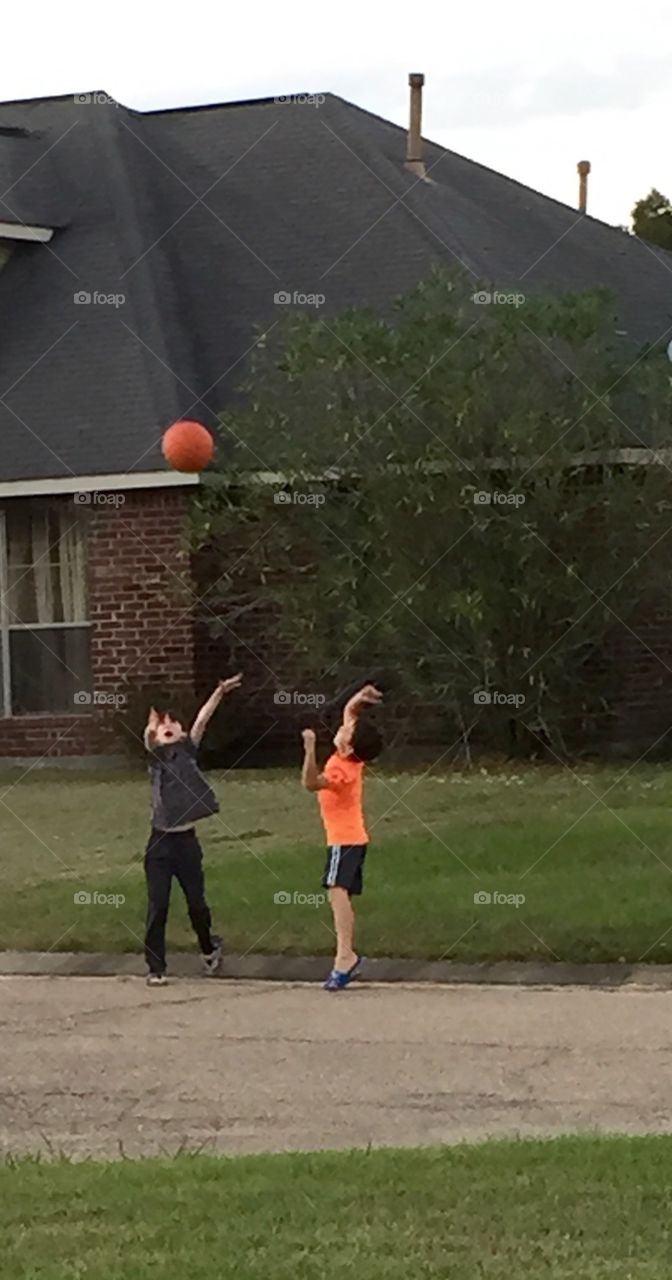 Two young boys playing basketball outside ,right by house. Having fun with arms raised and ball in the air.