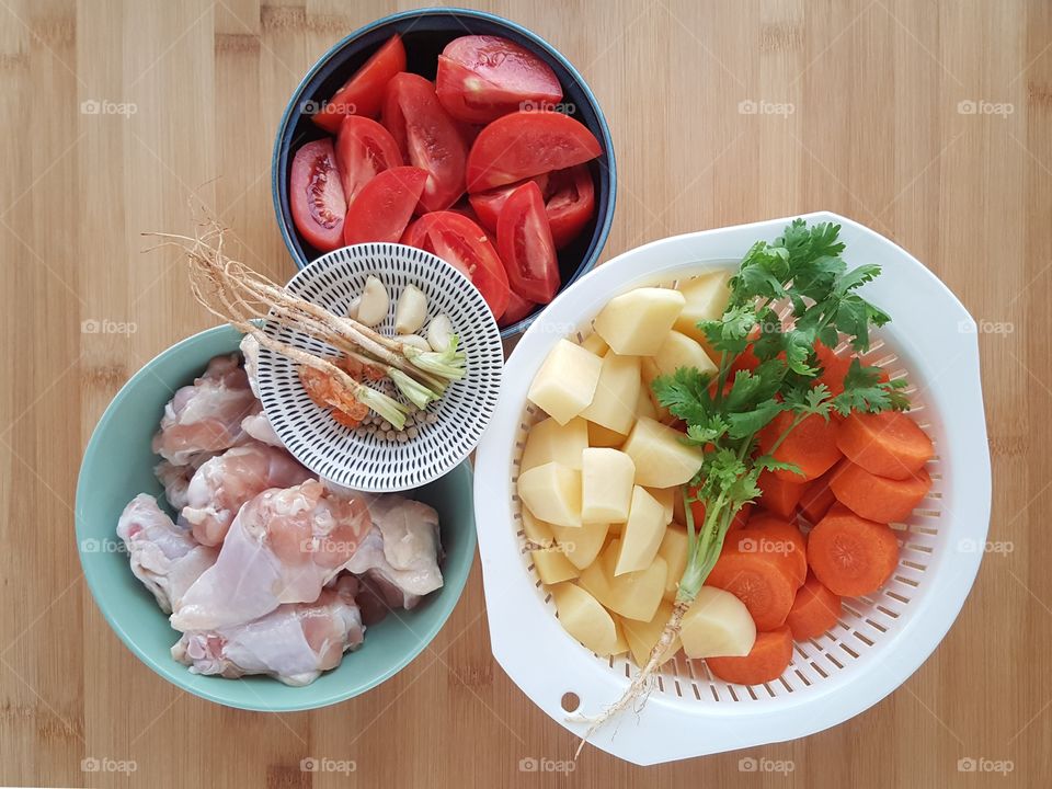The ingredients being prepared for cooking Asian cuisine -  cube potato and rondelles carrots in the white plastic colander, fresh chicken top wing, sliced tomatoes and coriander leaves. -  Close up