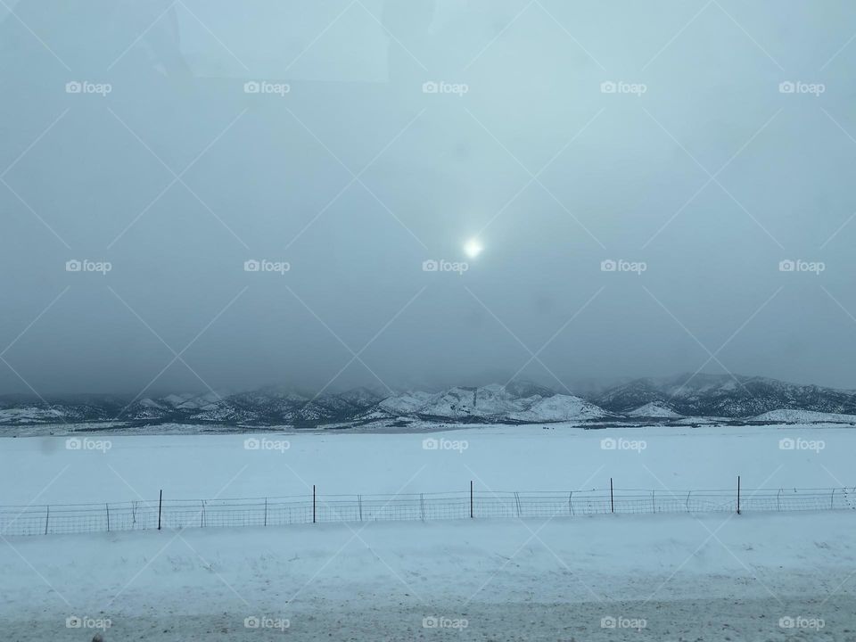 A snowy field with snowy mountains behind it and the sun above.