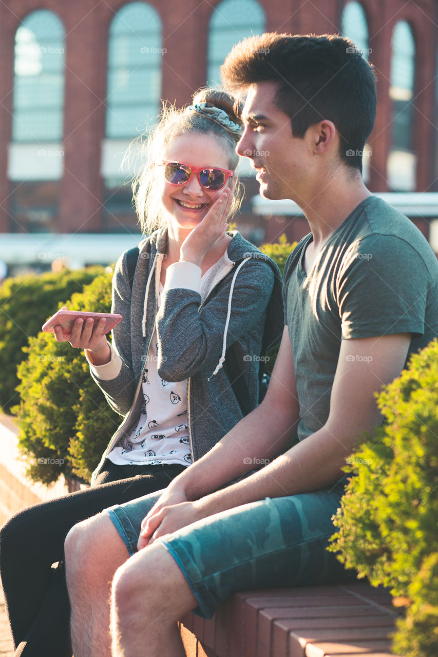 Couple of friends, teenage girl and boy,  having fun together, using smartphones,  sitting in center of town, spending time together. Real people, authentic situations