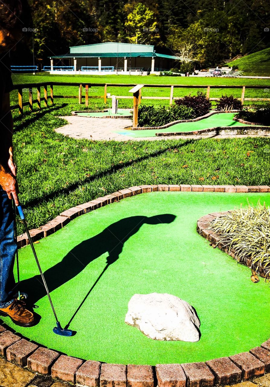 Person playing mini golf with sun behind them casting shadow on green