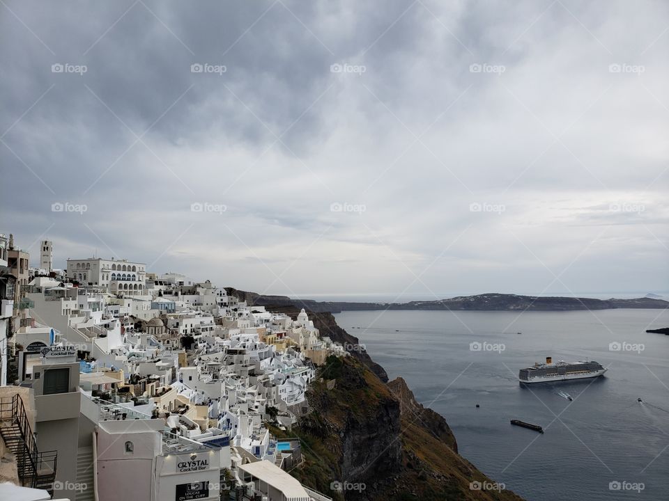 Side view from the height of Santorini, an island in Greece, with the white and yellow buildings on the cliff side and the cruise ship and boats in the Aegean sea below.