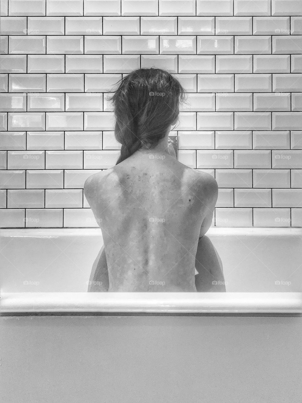 Naked woman sitting with her back to the camera against metro tiles in a bath