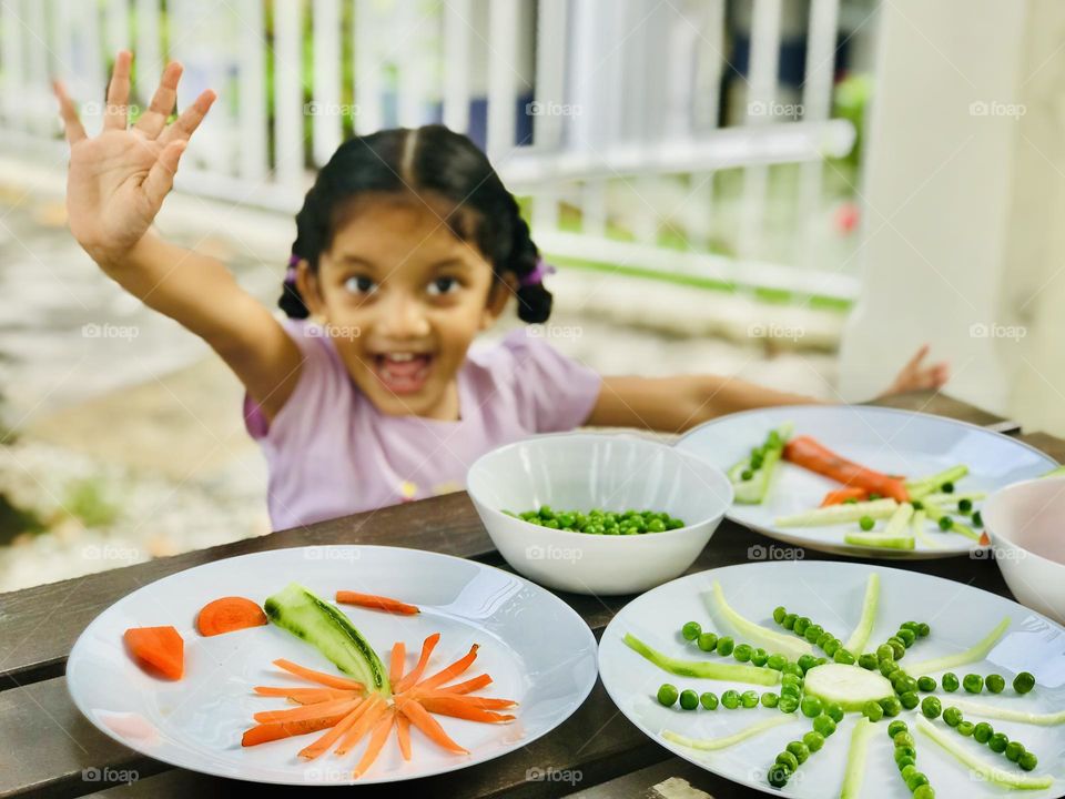 A four year old girl smiling and enjoying by doing creative craft coconut tree and sun with vegetables like carrots and cucumbers and Greenpeace.