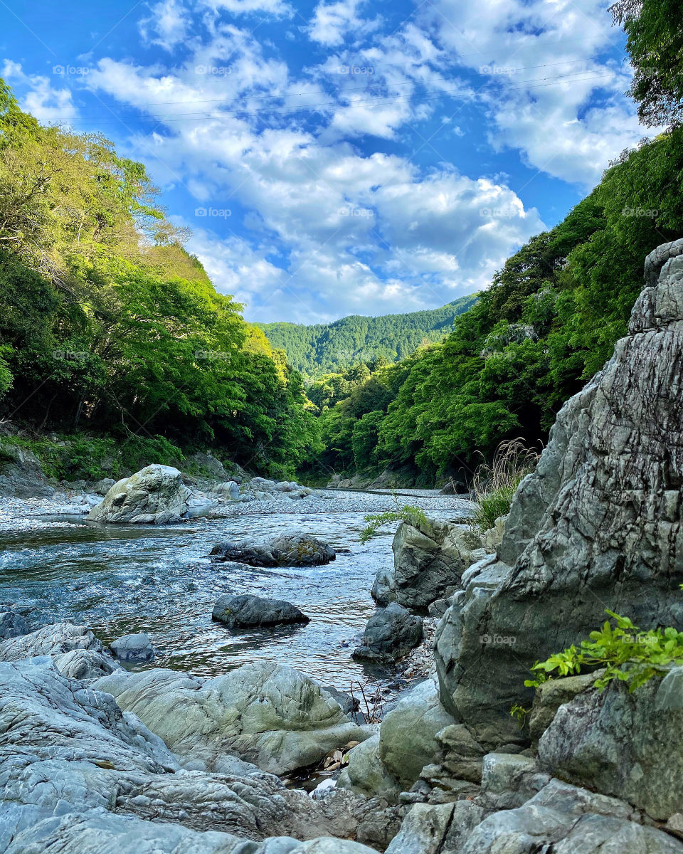 Nature scene of blue sky and fluffy white clouds above a flowing river with large gnarly rocks and surrounded by trees. 
