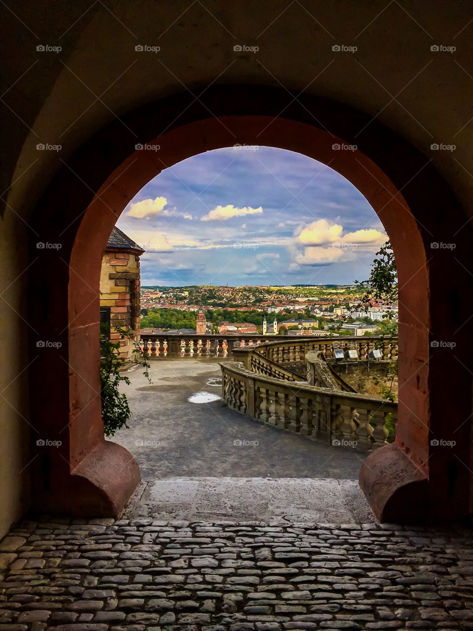 A stone built arch at the end of a cobblestone walkway; overlooking a colorful city under a blue-sky summer day.