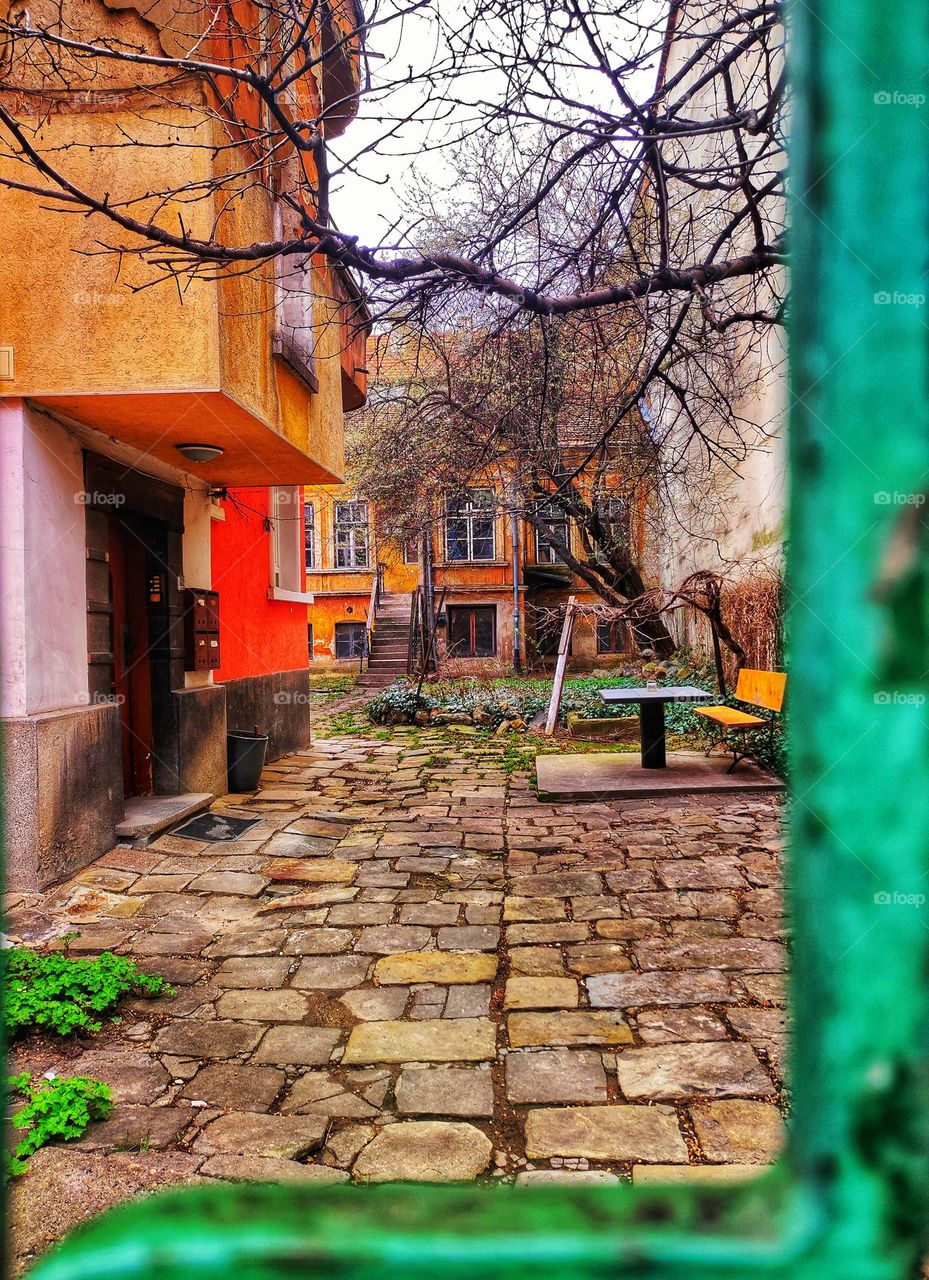 A beautiful photo of old building's back yard in the capital of Bulgaria with vibrant colours, trees, pavement and a green fence