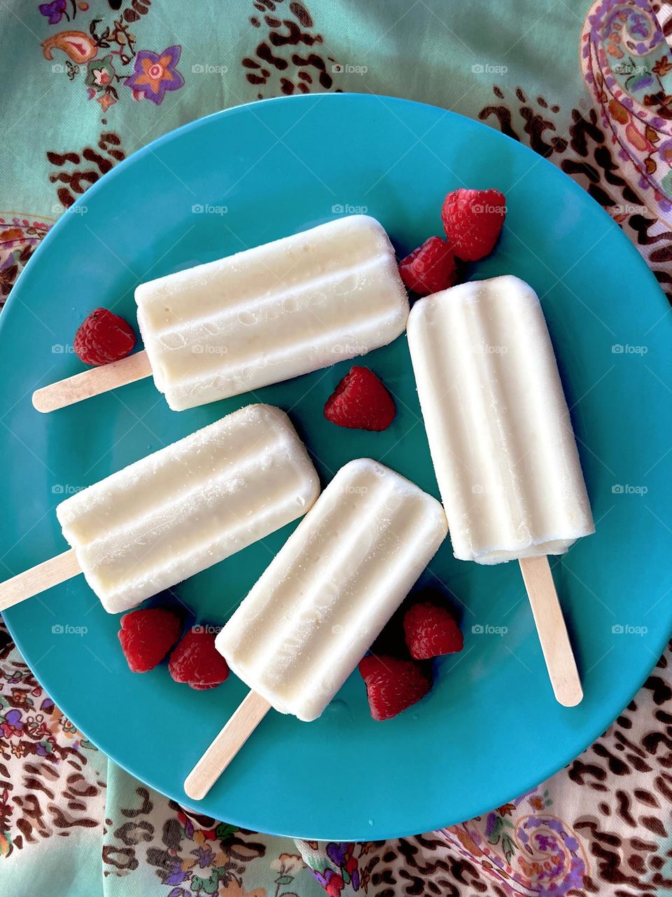 Four white coconut flavored popsicles with red raspberries on a turquoise blue plate set on a multicolored printed scarf