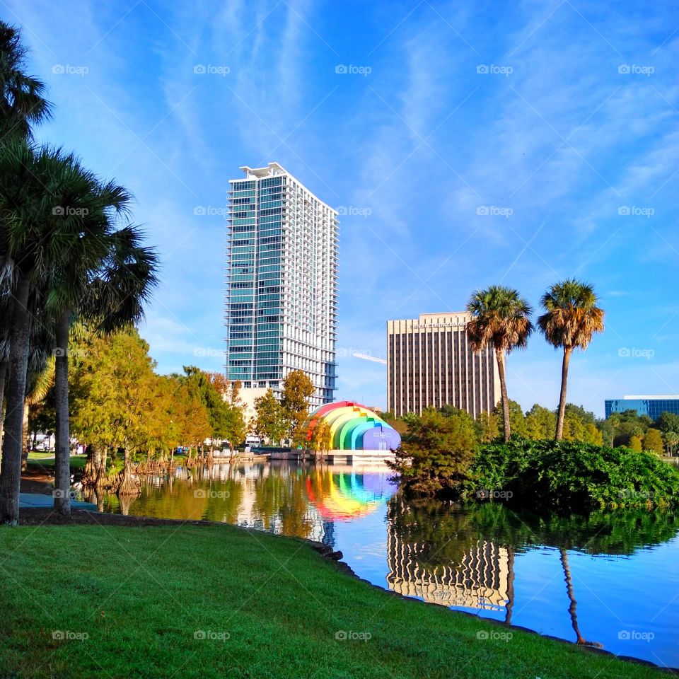 A landscape/cityscape photo of Lake Eola, and the surrounding land, including the rainbow amphitheater and city buildings. Also shown are palm trees.