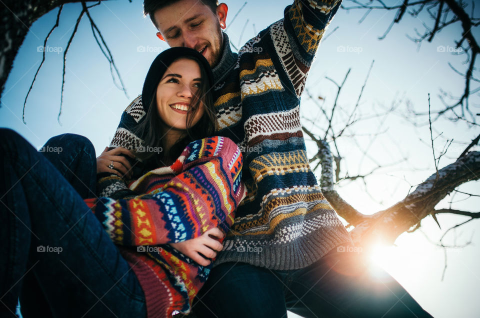 Joyful cute couple embraces. Young man in a sweater hugs a girl from sitting on tree branch in winter.
