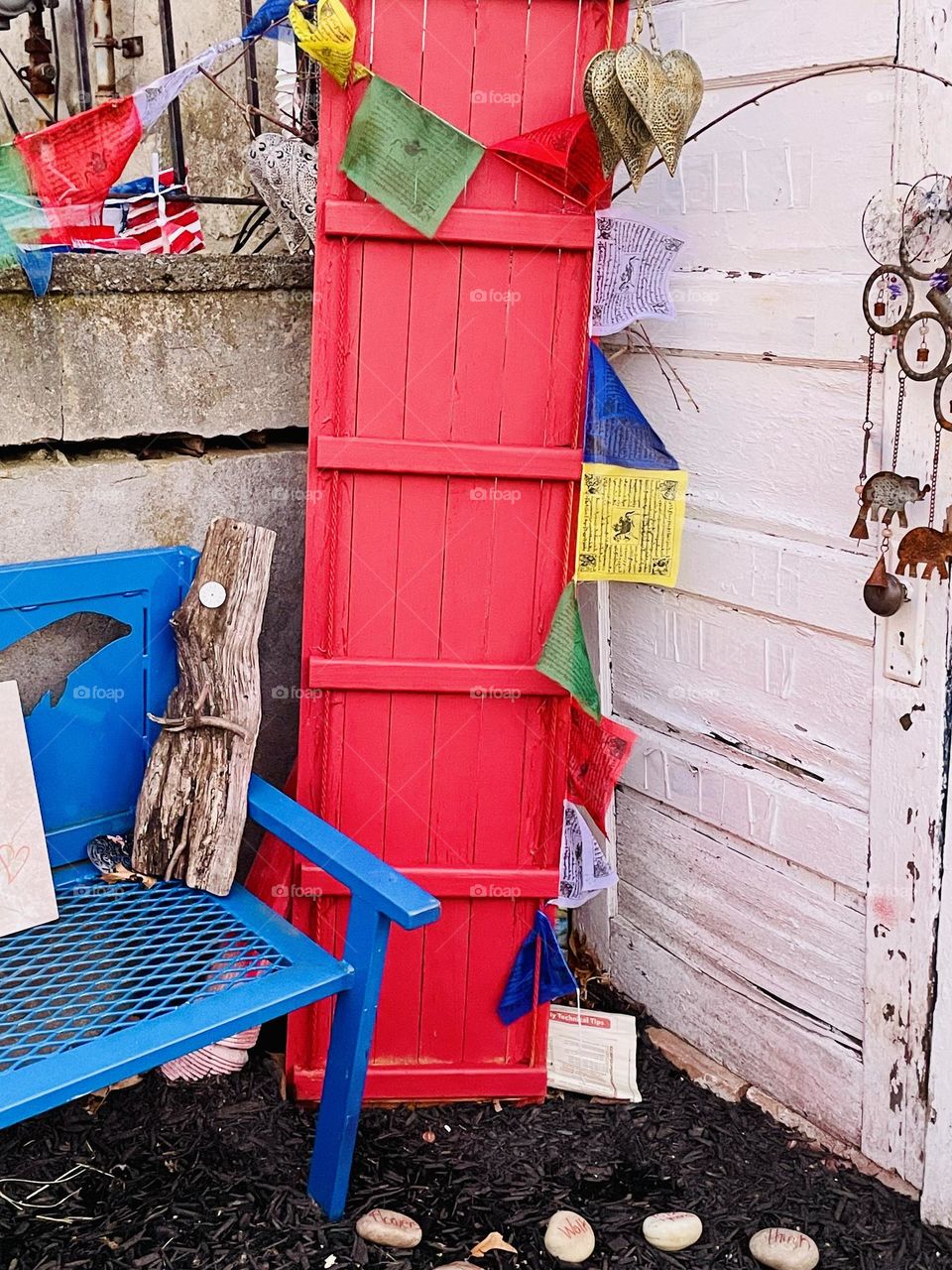 A weathered white door meets a bold red toboggan, propped casually beside it, draped in colorful flags—navy, white, yellow, green, red. A deep blue bench sits nearby surrounded by eclectic trinkets in a vibrant outdoor display.