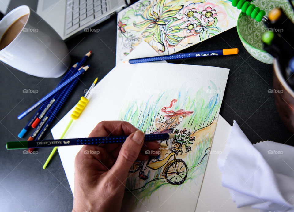 Closeup of hand holding Faber-Castell Aquarelle Colored watercolor pencils sketching girl on bicycle in countryside with sketchbook on table with laptop and coffee conceptual work life balance, creativity, hobby artist lifestyle photography