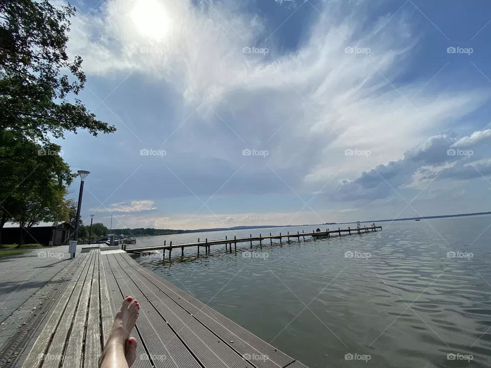 Weitwinkelaufnahme eines Stegs am Steinhuder Meer mit Schlierwolken und leicht verdeckter Sonne. Im unteren Bildteil sind zwei schöne Füße zu sehen/   