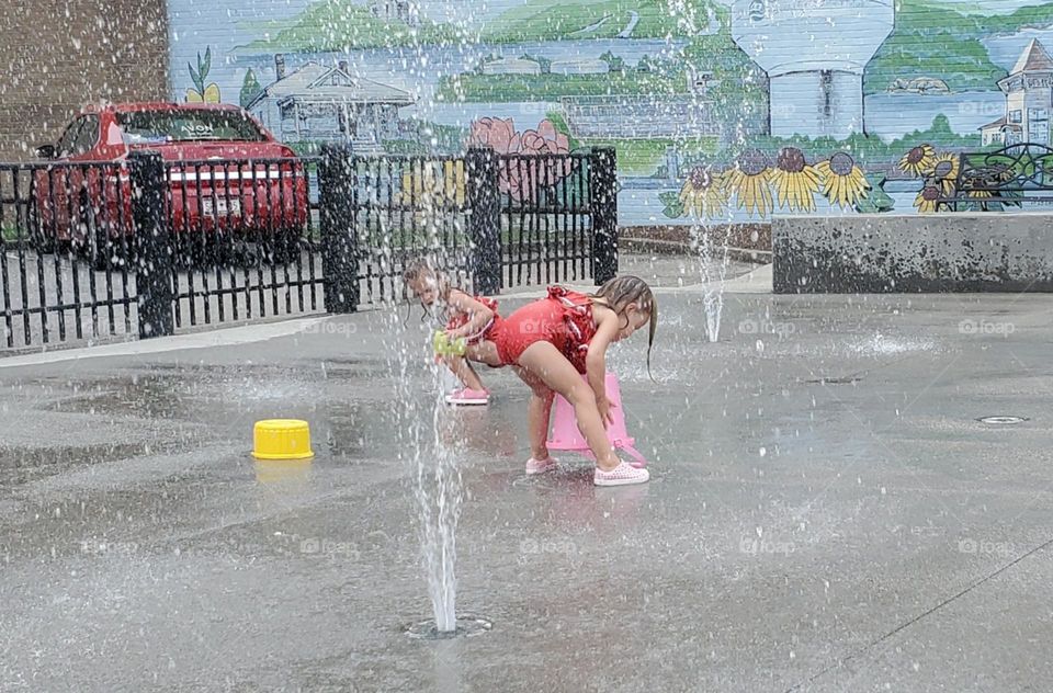 two little girls in red swimsuits playing in the splash pad in the city with buckets