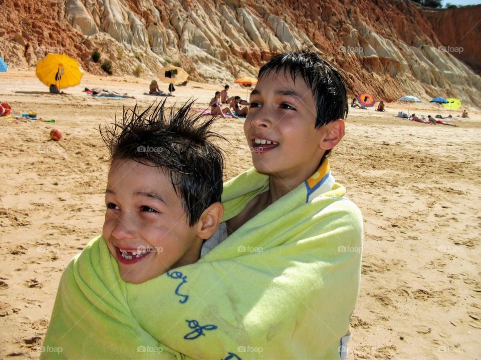 Two young boys at the beach, wrapped in the same green beach towel, smiling broadly at some joke their father told.