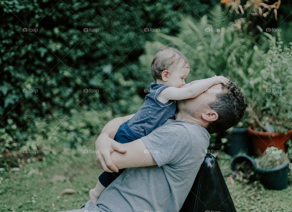 Handsome young caucasian father in a shirt with brown curly hair plays hugging with his little daughter while sitting on a long black garden chair in the backyard garden after work, side view closeup. The concept of fathers, love dad, gentle hugs, ou