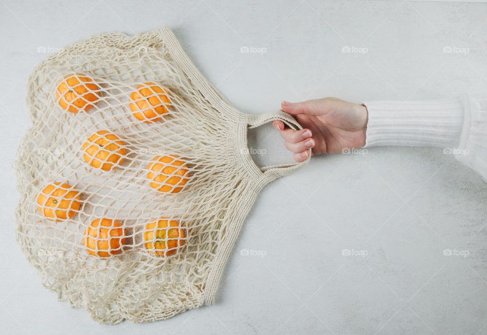 The hand of a young Caucasian girl in a white sweater holds a koton net with tangerines in it against a light cement background, side view. The concept of healthy food, tropical fruits.