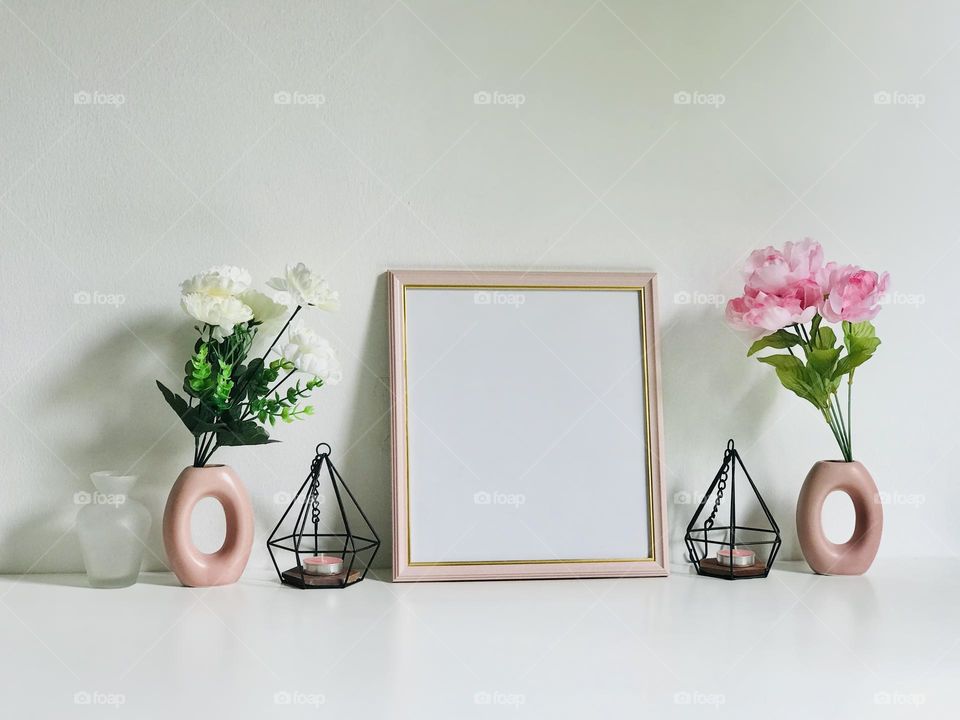 A pink colour photo frame along with pink and white flowers in pink flowers vases and pink candle in candle holder on white table looks pretty π