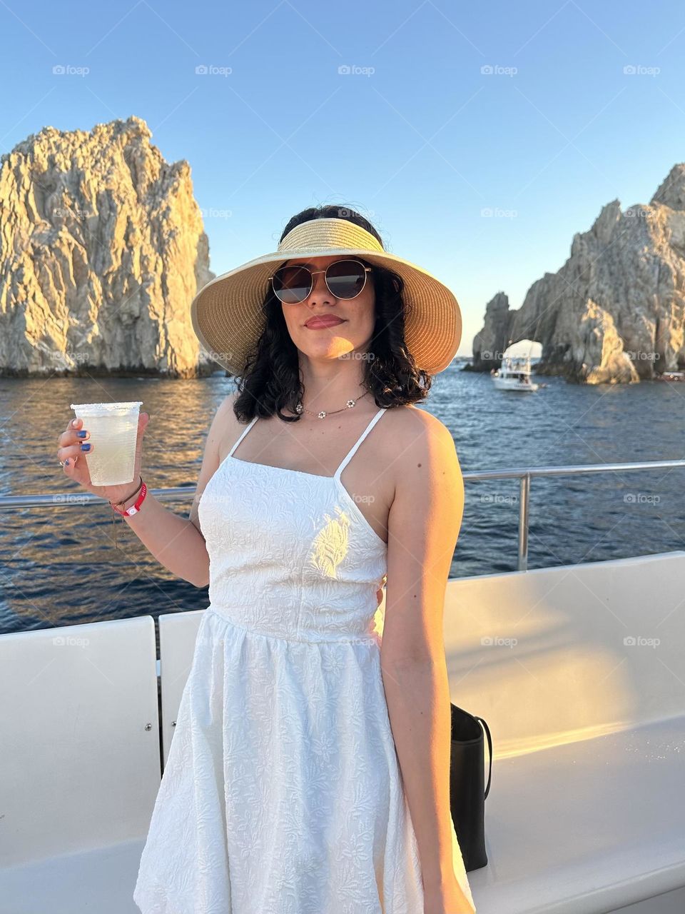 Woman wearing a hat and sunglasses, while holding a glass in her hand, standing on a boat.