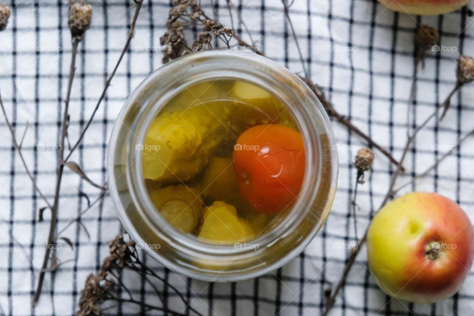 Flat lay of opened glass jar with pickled vegetables with a checkered napkin and apples on the background