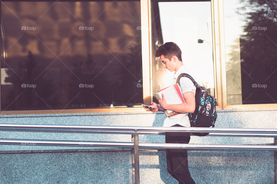 Student using mobile phone holding a notebook and carrying a backpack walking at the front of university building. Young boy wearing blue shirt and dark jeans