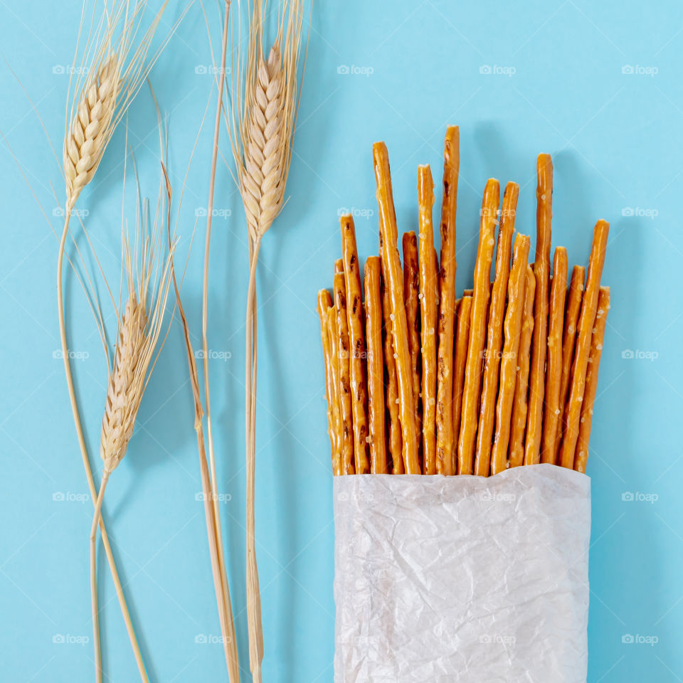 Flour snack. Salted, baked, wheat sticks in a bag made of white ecological paper with wheat spikelets on a blue background. Bread festival. Concept of natural products. Food photos, top view