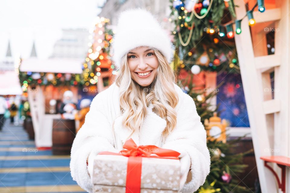 Young happy woman blonde girl with curly hair in white fur hat with gift box in hands at the Christmas fair market in winter street decorated with lights