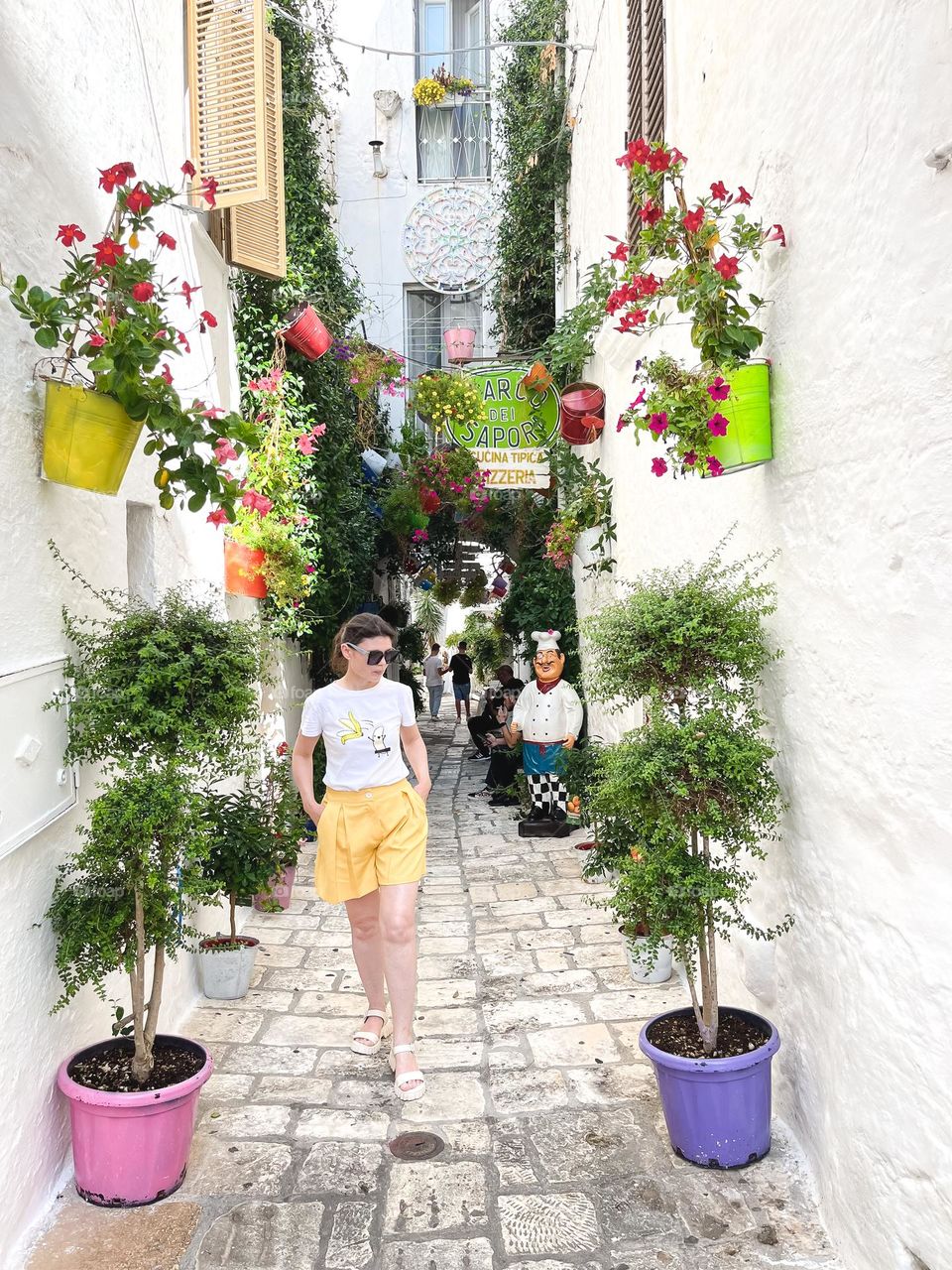 One young beautiful caucasian girl stands posing on a narrow white street with growing flowers in creative pots hanging on the walls and standing on the ground on a summer sunny day in Ostuni, Italy, close-up side view. Urban plants concept.